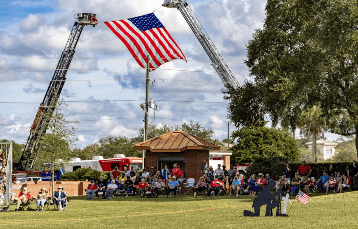 A large group of people gathers on a grassy field for a Memorial Day tribute. Two fire trucks extend ladders to display a giant American flag high in the sky. Attendees sit on lawn chairs facing a black silhouette statue of a kneeling soldier beside a cross and American flag, honoring fallen heroes. Trees, a small brick building, and a partly cloudy sky complete the backdrop.