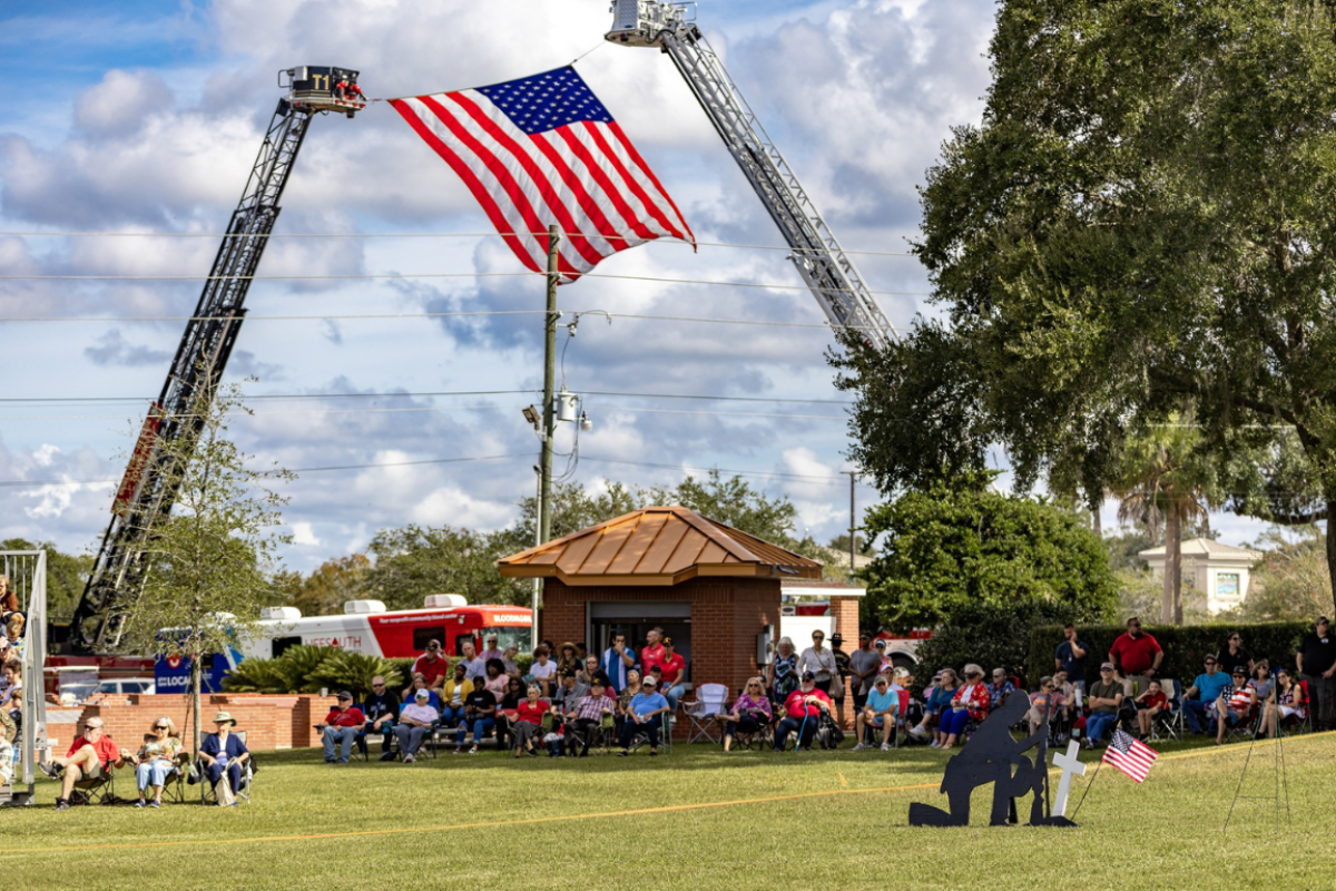 A large group of people gathers on a grassy field for a Memorial Day tribute. Two fire trucks extend ladders to display a giant American flag high in the sky. Attendees sit on lawn chairs facing a black silhouette statue of a kneeling soldier beside a cross and American flag, honoring fallen heroes. Trees, a small brick building, and a partly cloudy sky complete the backdrop.
