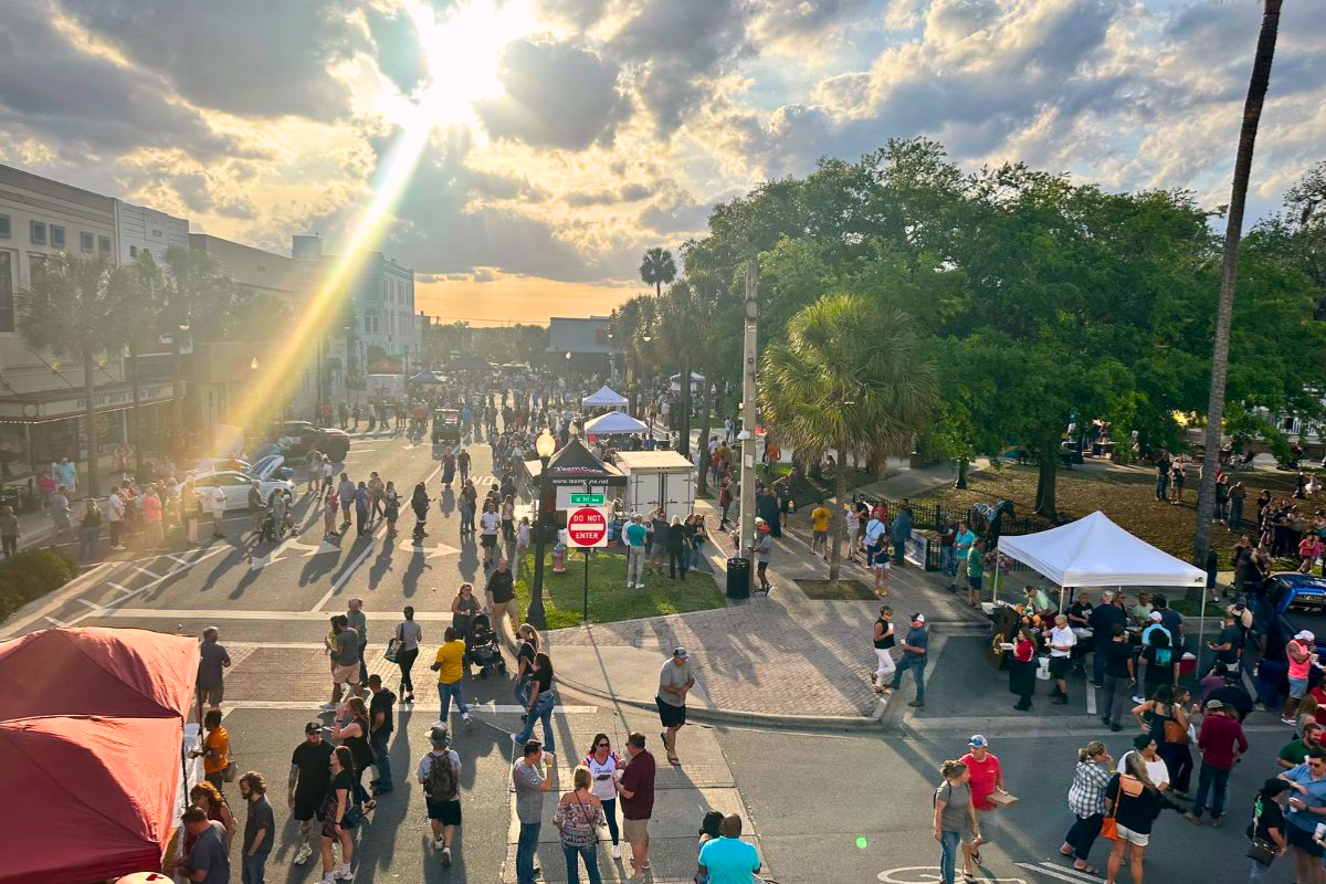 A lively downtown street festival at sunset with crowds of people walking, browsing vendor booths, and enjoying the event. The sun is shining through clouds, casting a warm golden glow over the scene. Palm trees, historic buildings, and white tents line the streets.