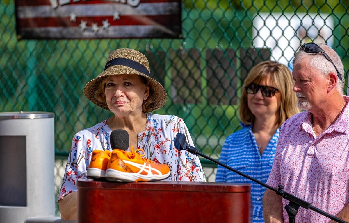 Patti Hunt, wearing a sun hat and floral blouse, speaks at a podium with a pair of orange athletic shoes in front of her and family members standing behind.