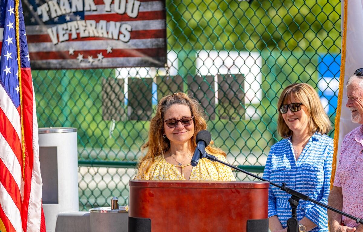 Angela, wearing sunglasses and a yellow top, speaks at a podium in front of a chain-link fence and American flag-themed "Thank You Veterans" sign.
