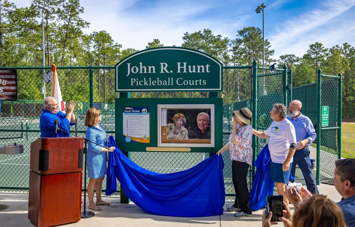 People unveiling a green sign reading “John R. Hunt Pickleball Courts” at Coehadjoe Park, with a photo of Hunt and his wife displayed beneath it.