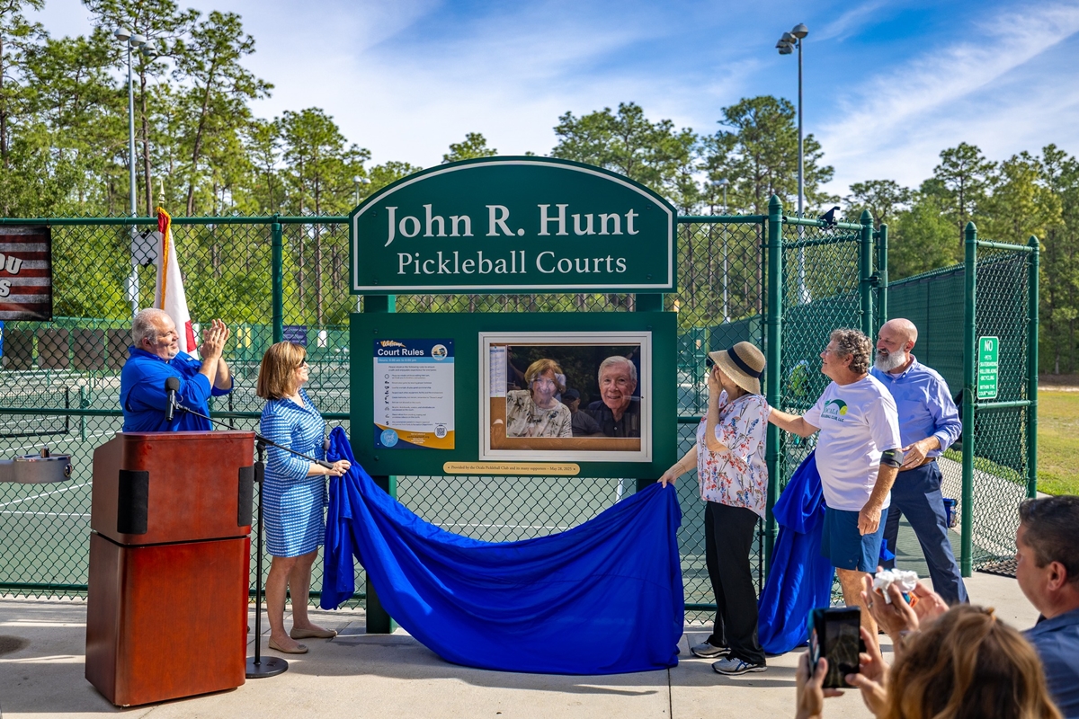 People unveiling a green sign reading “John R. Hunt Pickleball Courts” at Coehadjoe Park, with a photo of Hunt and his wife displayed beneath it.