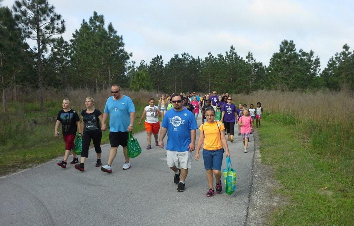 A large group of people of all ages walk along a paved trail surrounded by trees and grass during a RE/MAX 5K fundraiser at Baseline Road Trailhead Park.