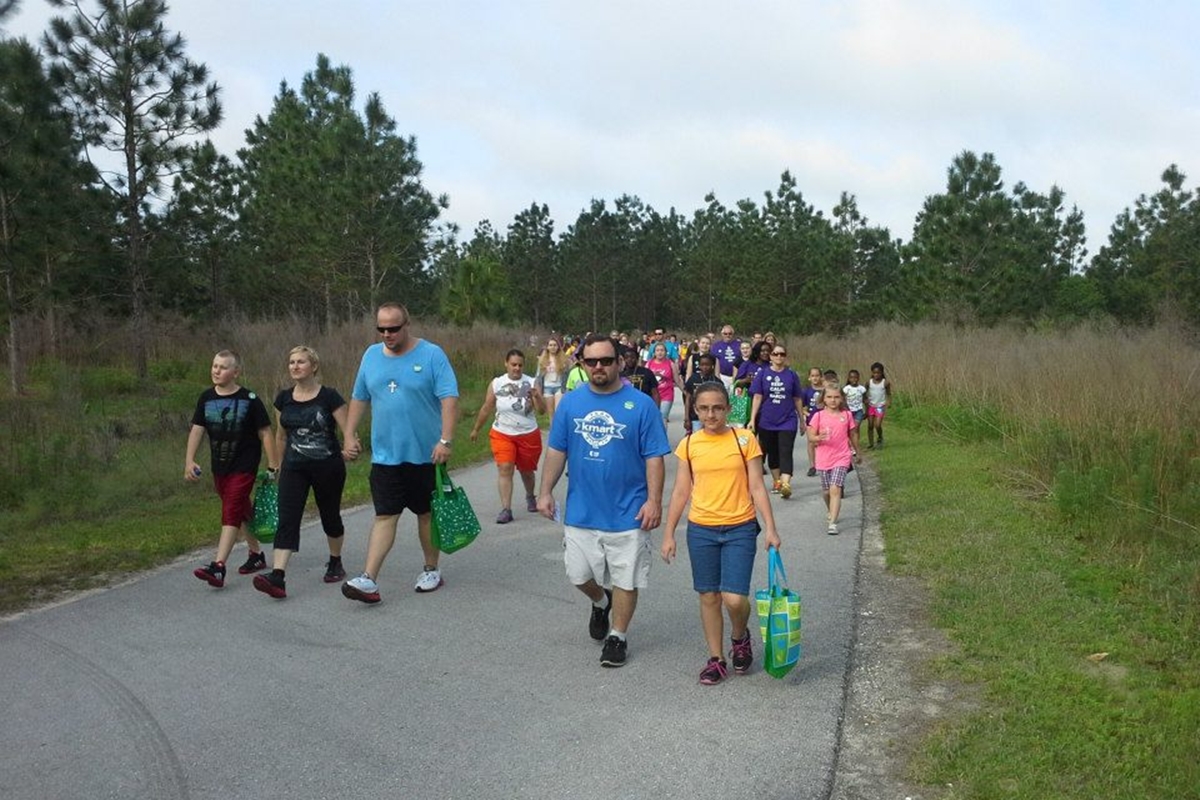 A large group of people of all ages walk along a paved trail surrounded by trees and grass during a RE/MAX 5K fundraiser at Baseline Road Trailhead Park.