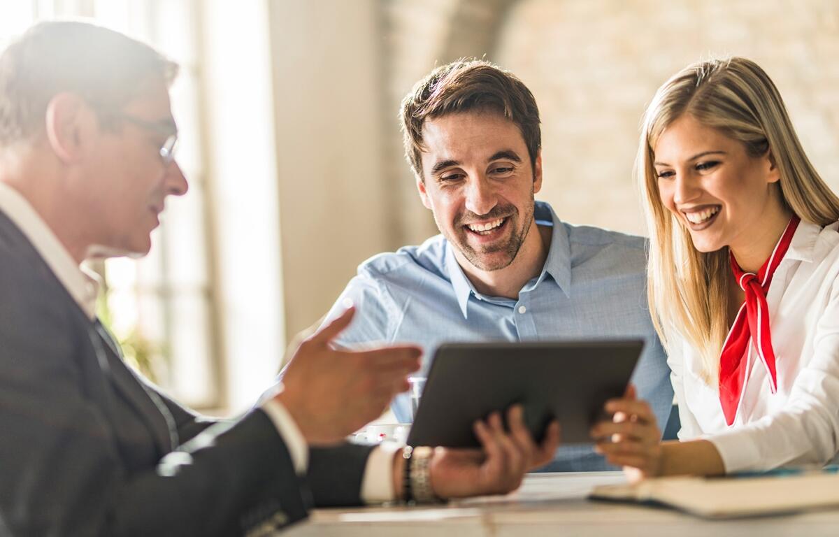 A smiling couple meets with a bank advisor, reviewing information on a tablet during a financial consultation.