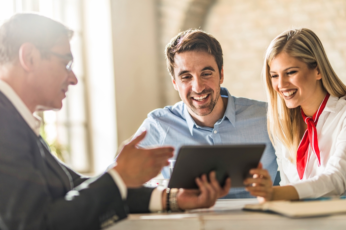 A smiling couple meets with a bank advisor, reviewing information on a tablet during a financial consultation.