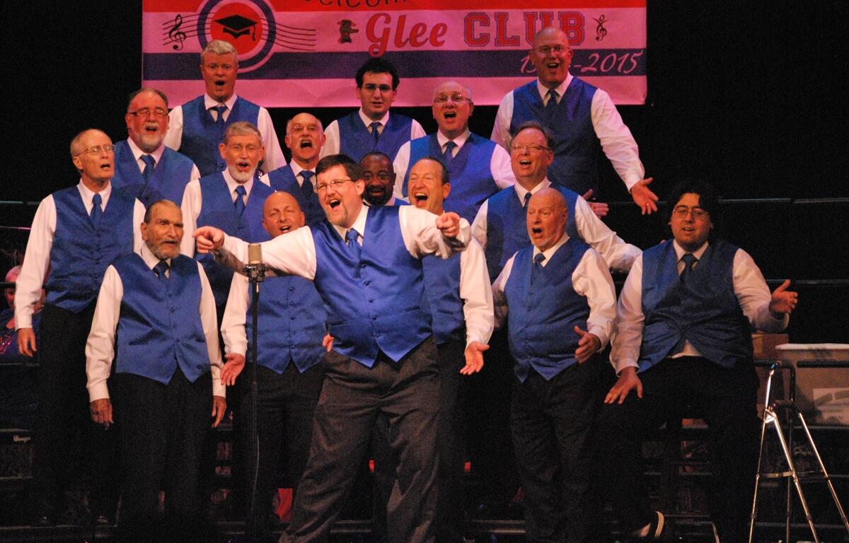 Barbergators Chorus members wearing matching blue vests sing together on stage in an energetic group performance.