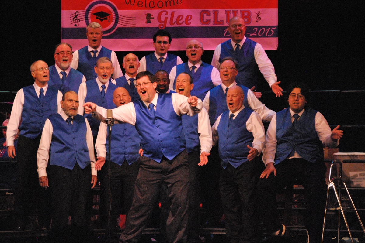 Barbergators Chorus members wearing matching blue vests sing together on stage in an energetic group performance.