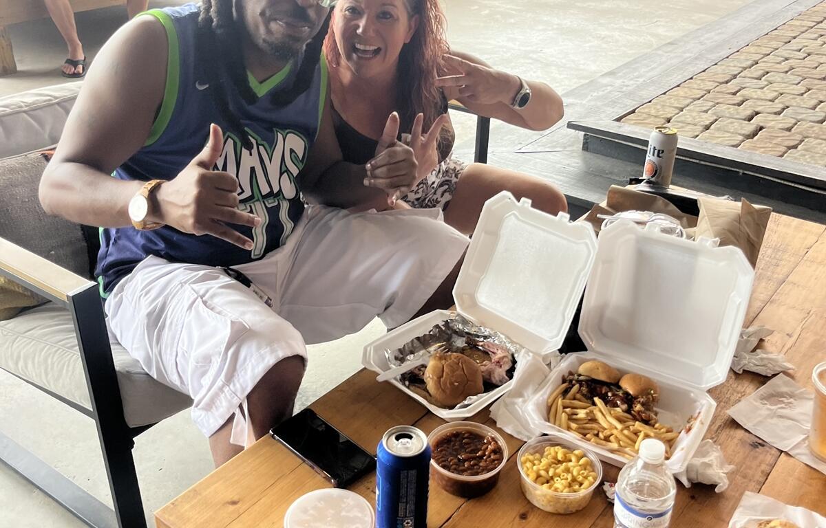 Man and woman smiling with plates of barbecue, fries, beans, and drinks at a festival table in Williston