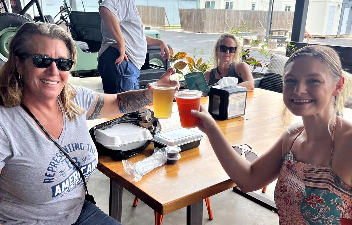 Two women smiling and clinking plastic cups of beer at a festival table with food containers nearby in Williston