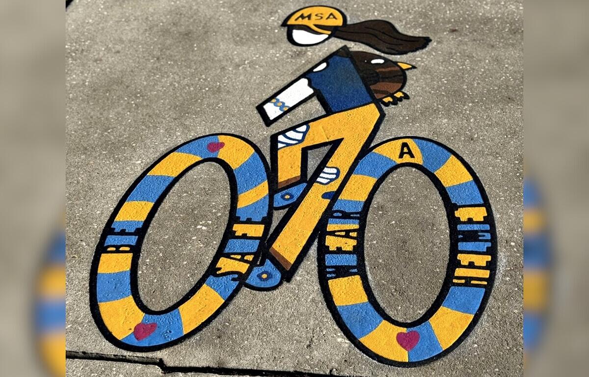 A colorful sidewalk mural of a cyclist, featuring bold yellow and blue stripes, hearts, and safety-themed words, painted as part of FDOT’s Bike Lane Design Contest near Madison Street Academy.