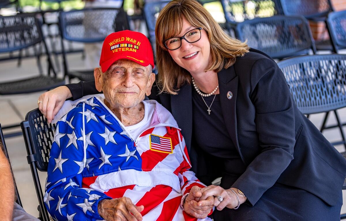 WWII veteran Howard Mautner wearing a patriotic jacket sits beside Commissioner Kathy Bryant, both smiling and holding hands at a Memorial Day event.