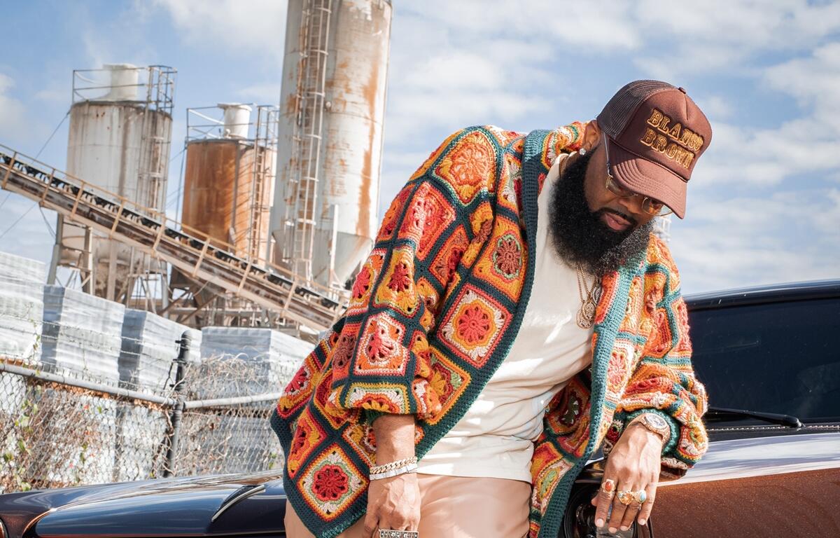 Blanco Brown leans on a classic brown car while wearing a colorful crocheted jacket, beige pants, and a brown cap with his name embroidered. Industrial silos and a blue sky fill the background.