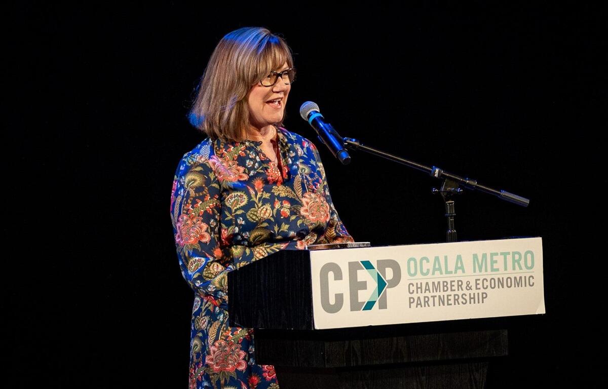 Kathy Bryant stands at a podium with an Ocala Metro Chamber and Economic Partnership sign, speaking into a microphone during the State of the County event.