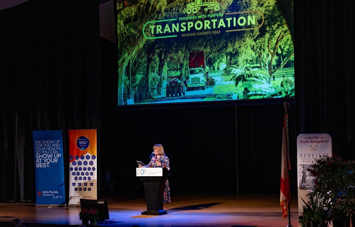 Kathy Bryant speaks at a podium with a large screen behind her reading “Transportation: Marion County 2025,” featuring an image of trucks on a tree-lined road.