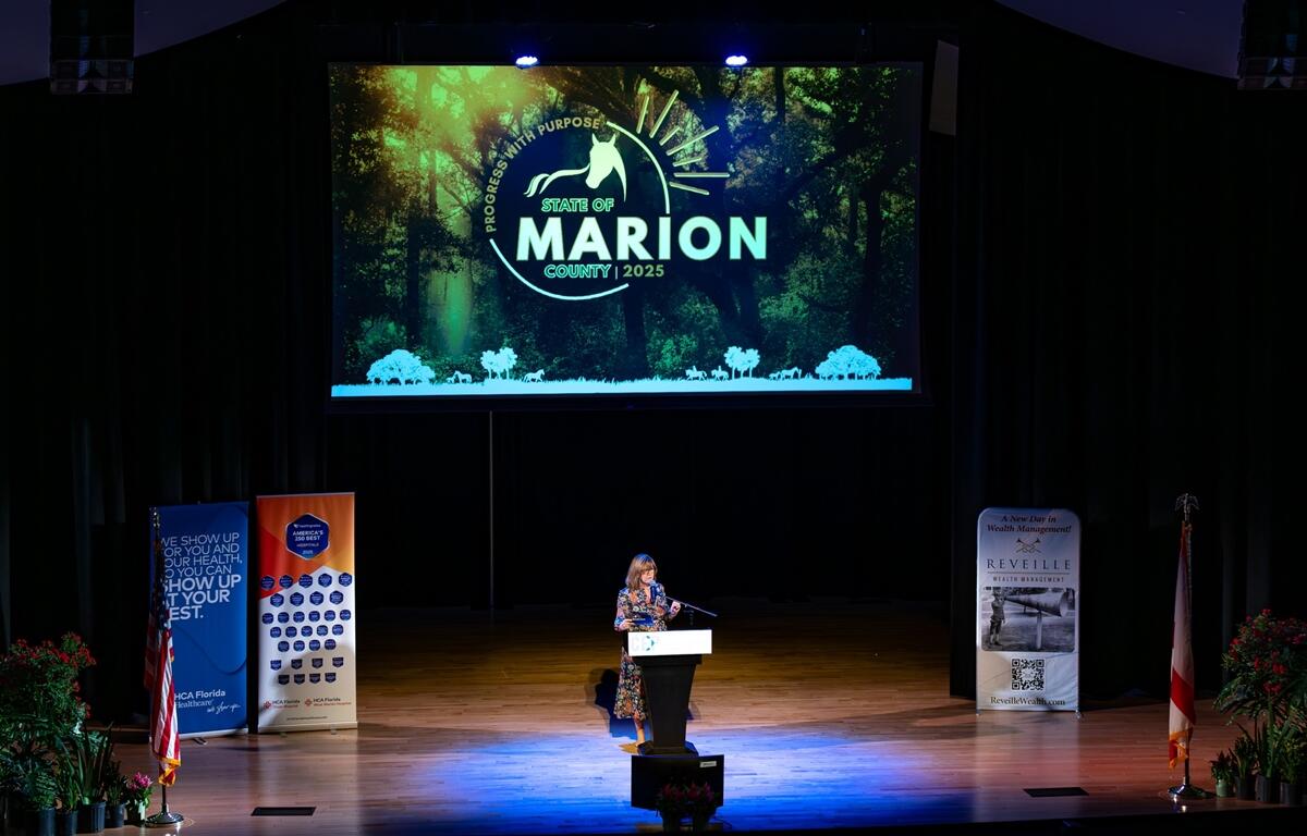 Kathy Bryant speaks at a podium under a large screen reading "State of Marion County 2025" during the State of the County address at the Reilly Arts Center.