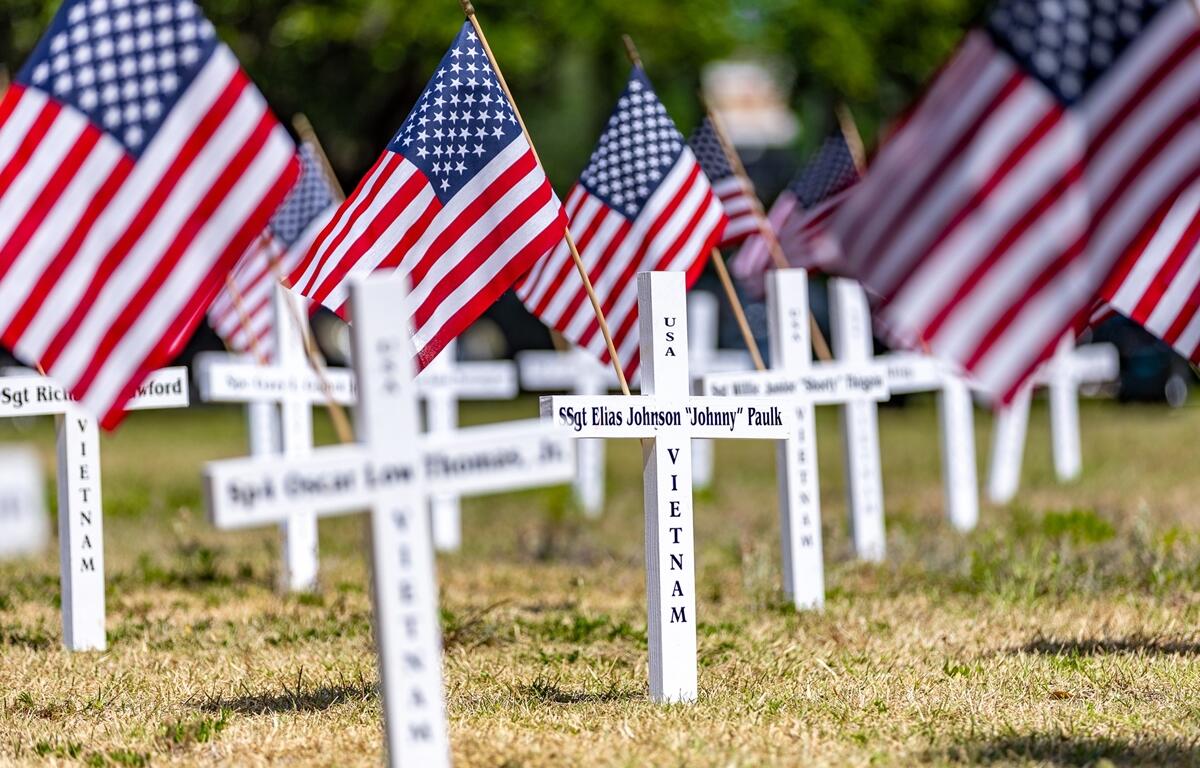 Rows of white crosses with names of Vietnam veterans and American flags are displayed on grass as a tribute during a Memorial Day event.