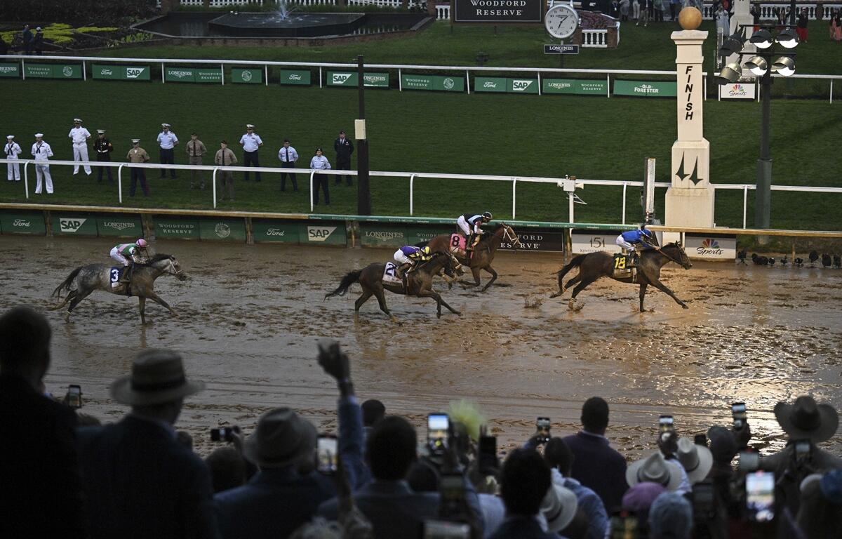 Racehorse Sovereignty, wearing number 14 and ridden by a jockey in blue silks, crosses the finish line first on a muddy track at Churchill Downs during the 151st Kentucky Derby. Several other horses trail closely behind as a crowd of spectators, many in hats, cheer and take photos from the stands. Uniformed personnel line the grassy infield near the iconic Derby finish post.