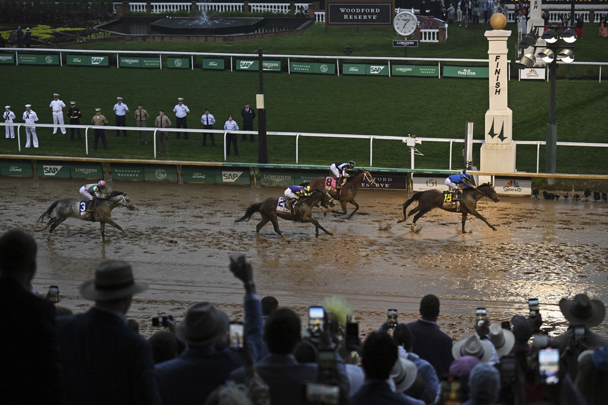 Racehorse Sovereignty, wearing number 14 and ridden by a jockey in blue silks, crosses the finish line first on a muddy track at Churchill Downs during the 151st Kentucky Derby. Several other horses trail closely behind as a crowd of spectators, many in hats, cheer and take photos from the stands. Uniformed personnel line the grassy infield near the iconic Derby finish post.