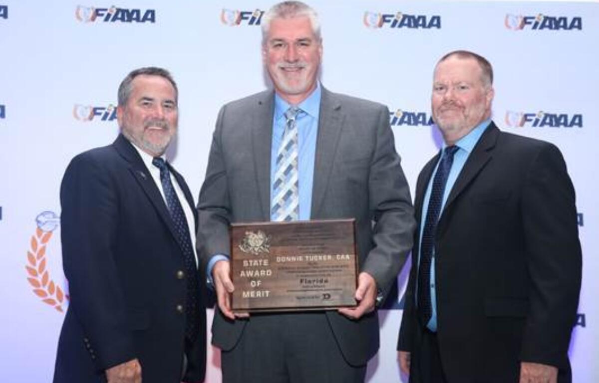 Three men in suits pose for a photo at the FIAAA conference in Kissimmee, Florida. Donnie Tucker, standing in the center, holds a plaque that reads "State Award of Merit."