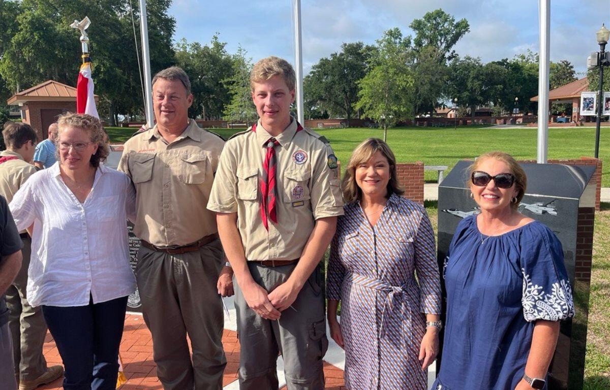 A woman in a white blouse, with the Flag of the United States of America, stands next to two men dressed in scout uniforms, who are standing next two women in dresses at the military service display flag plaza at the Ocala/Marion County Veterans Memorial Park.