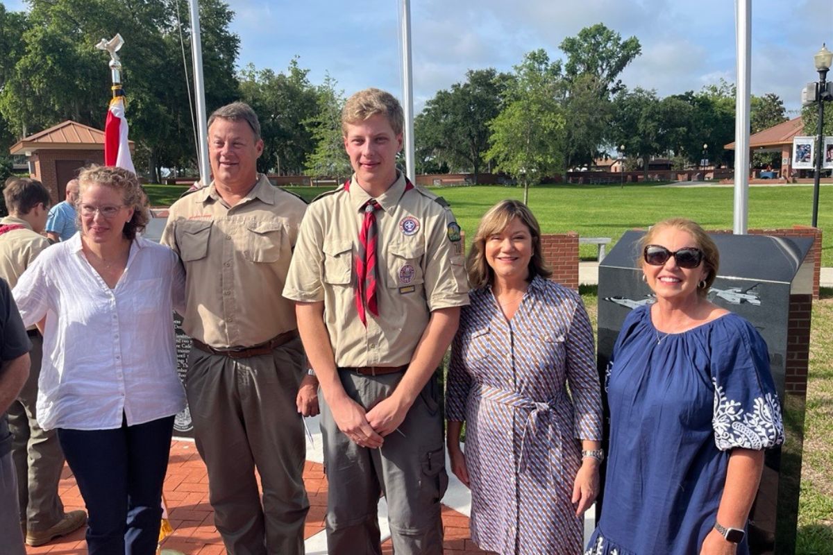 A woman in a white blouse, with the Flag of the United States of America, stands next to two men dressed in scout uniforms, who are standing next two women in dresses at the military service display flag plaza at the Ocala/Marion County Veterans Memorial Park.