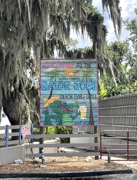 Colorful hand-painted sign reading “Gator Joe’s Beach Bar & Grill” featuring cartoon alligators and beach imagery, displayed near entrance with Spanish moss-covered trees in background.