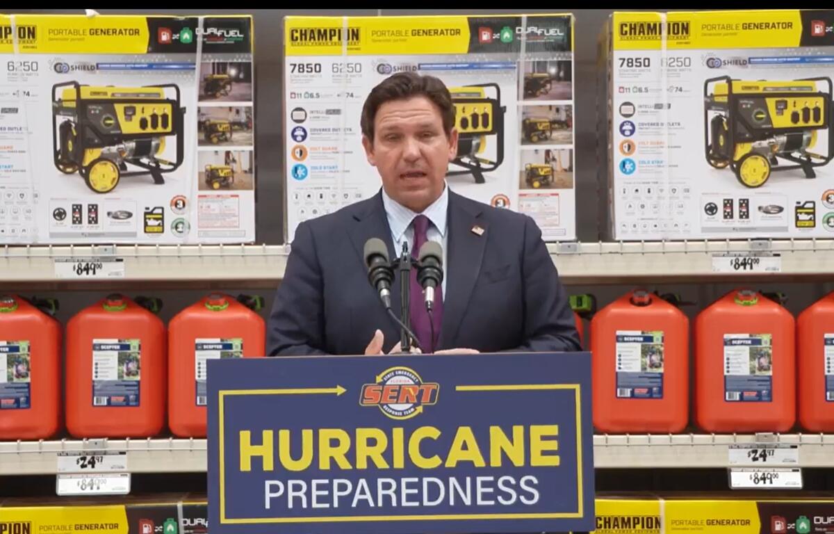 Florida Gov. Ron DeSantis stands at a podium labeled “Hurricane Preparedness” inside a Home Depot, with gas cans and portable generators on display behind him.