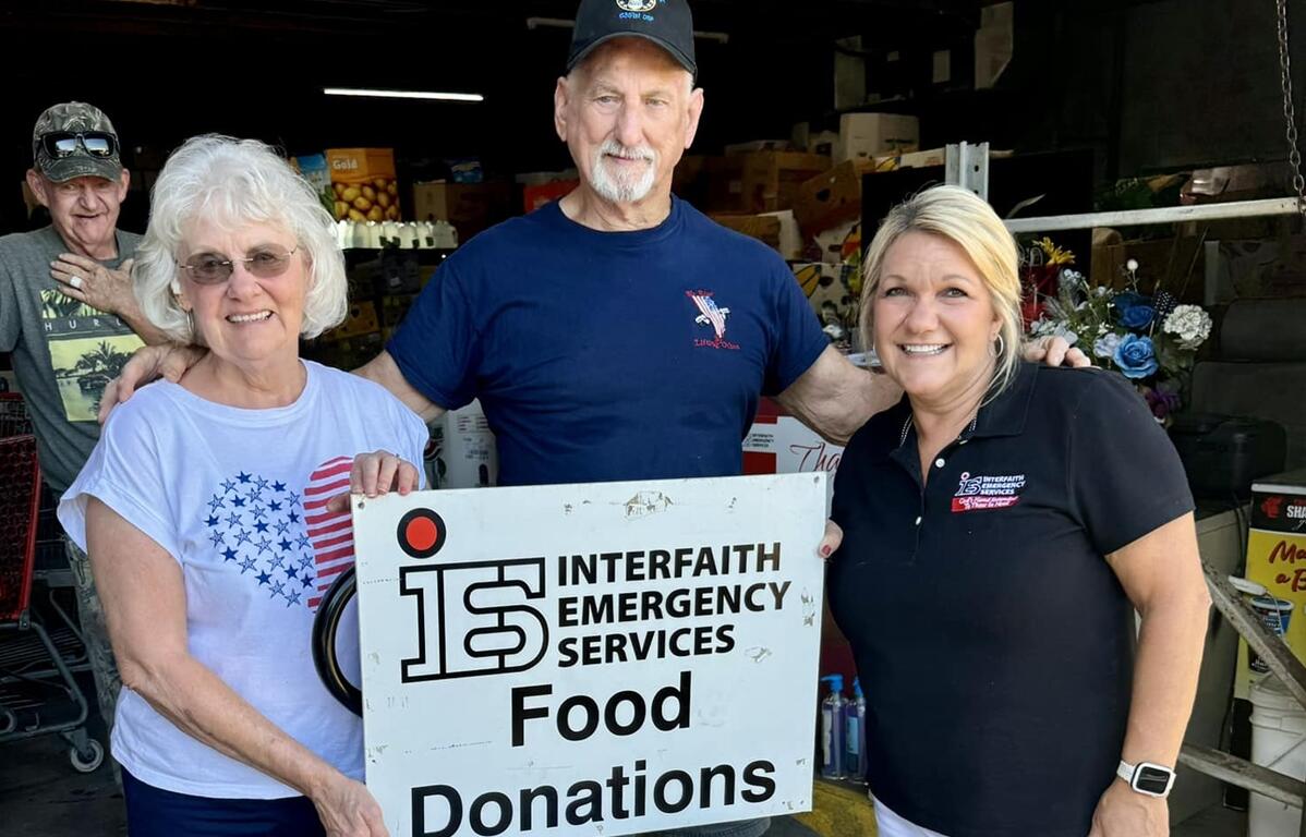 Karla Grimsley poses with two Twilight Cruisers members holding a sign that reads “Interfaith Emergency Services Food Donations,” with shelves of food items visible in the background.
