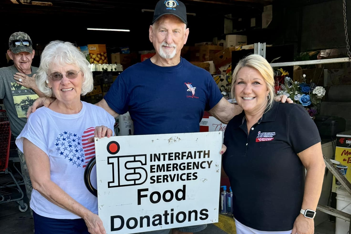 Karla Grimsley poses with two Twilight Cruisers members holding a sign that reads “Interfaith Emergency Services Food Donations,” with shelves of food items visible in the background.