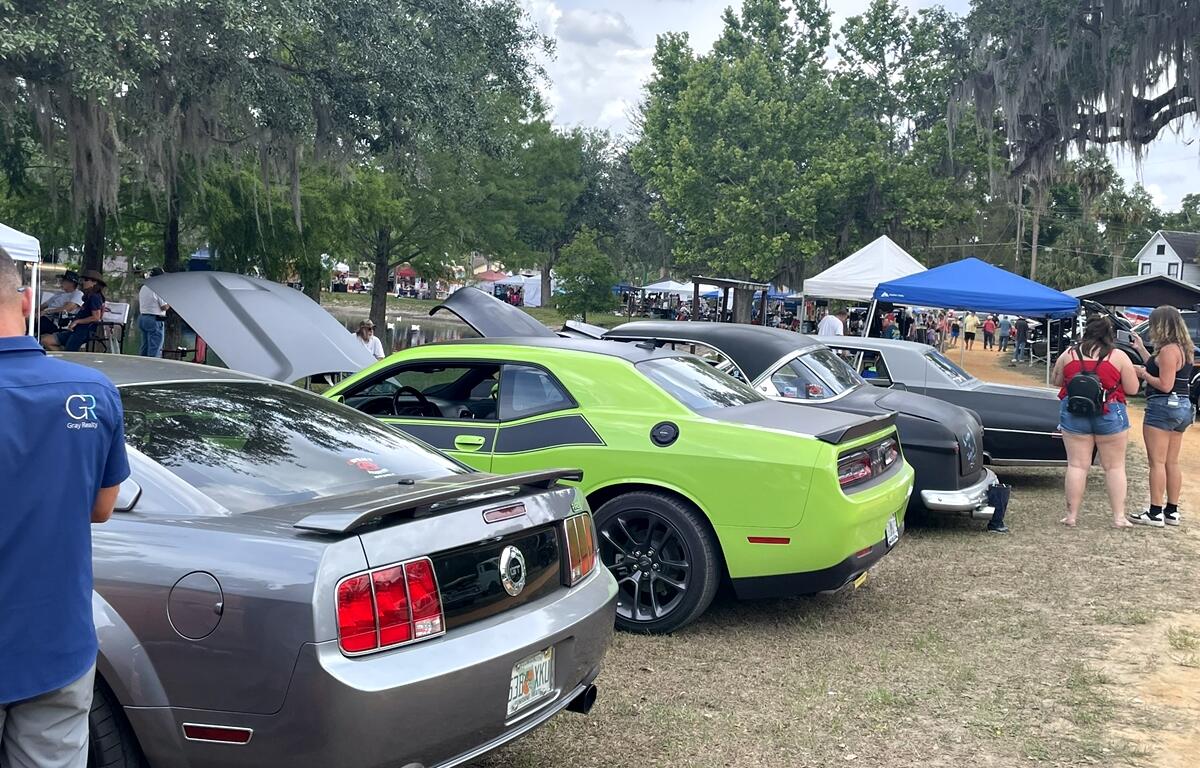 People walk beside parked muscle cars, including a bright green Dodge Challenger, at an outdoor car show with vendor tents and large trees in the background.