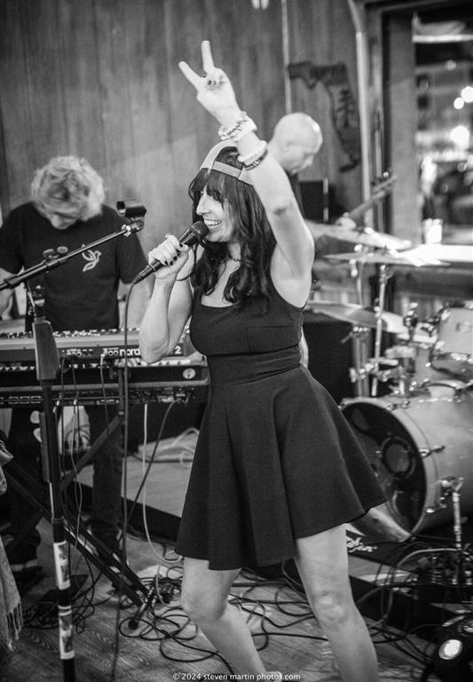 Heather Lynne sings on stage with her band, wearing a black dress and holding a microphone, as she raises one arm in a rock gesture.