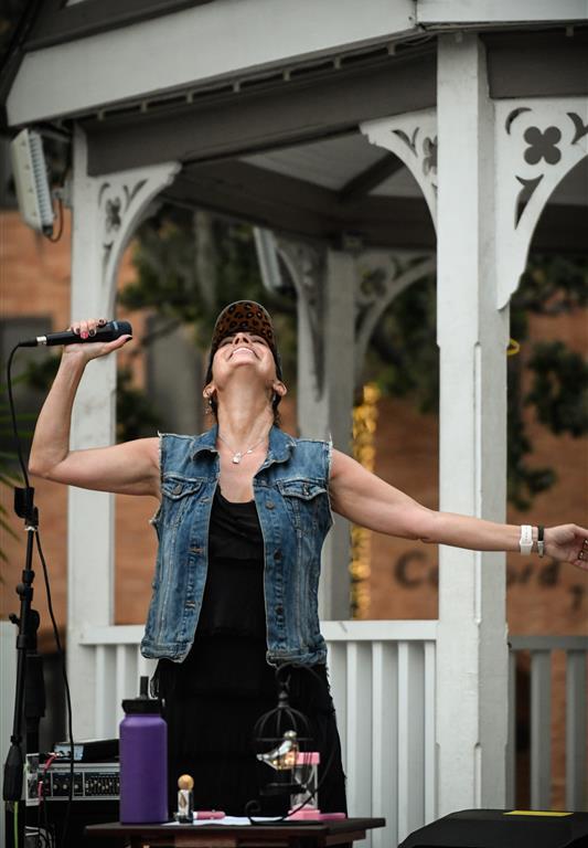 Heather Lynne stands in a gazebo with arms wide open and head tilted back, holding a microphone and wearing a denim vest and cap during a live outdoor show.