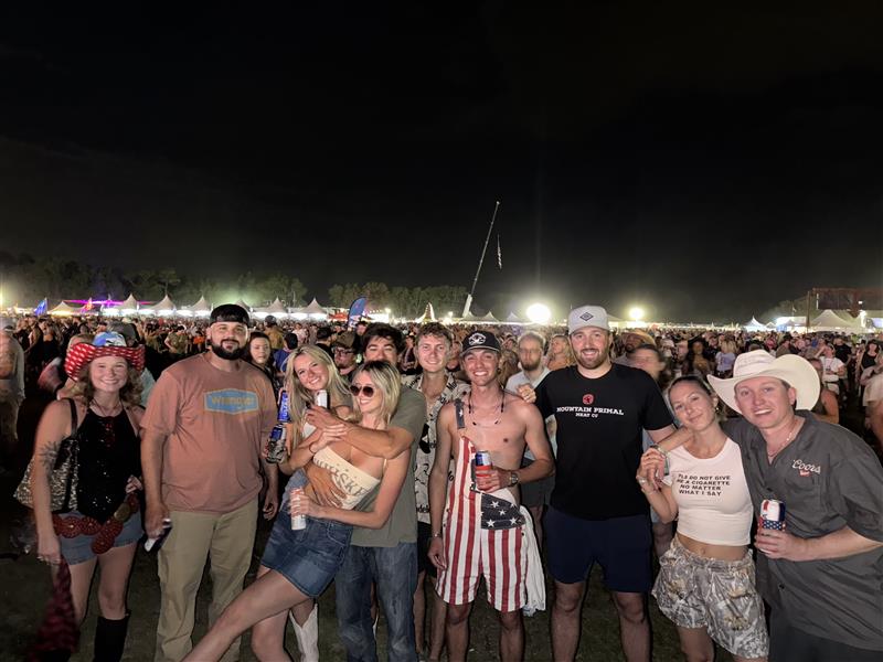 A group of young adults pose for a photo at night during the Rock the Country festival, with some dressed in patriotic-themed outfits and holding drinks, surrounded by a large crowd and tents glowing under festival lights in the background.