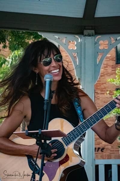 Heather Lynne plays acoustic guitar and sings into a microphone, smiling in sunglasses during a live outdoor performance at the gazebo in downtown Ocala.