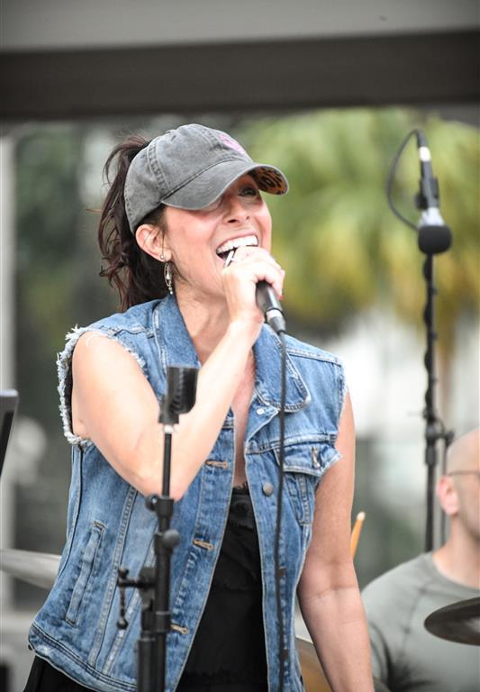 Heather Lynne sings into a microphone during an outdoor show, wearing a denim vest and baseball cap, with a drummer visible in the background.