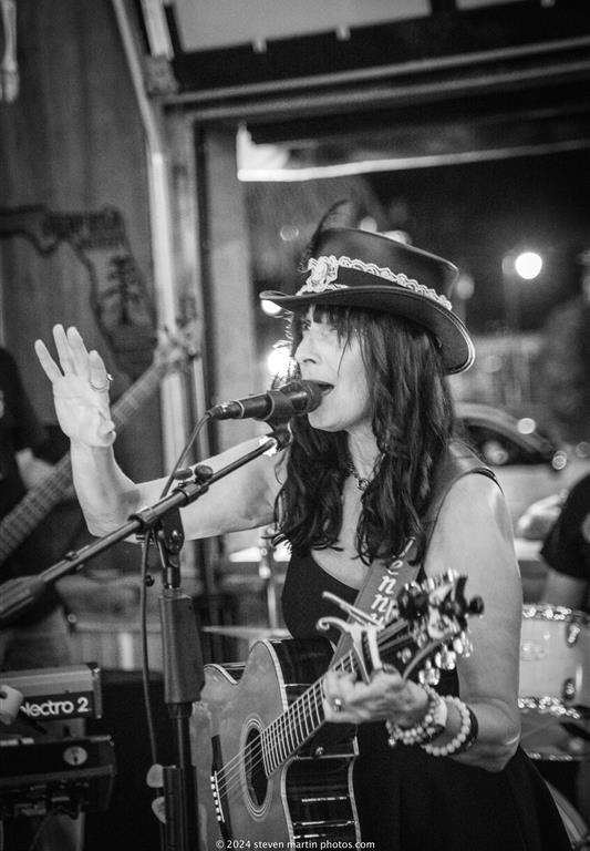 Heather Lynne performs with a guitar and sings into a microphone, raising one hand mid-song while wearing a feathered top hat during a live show.