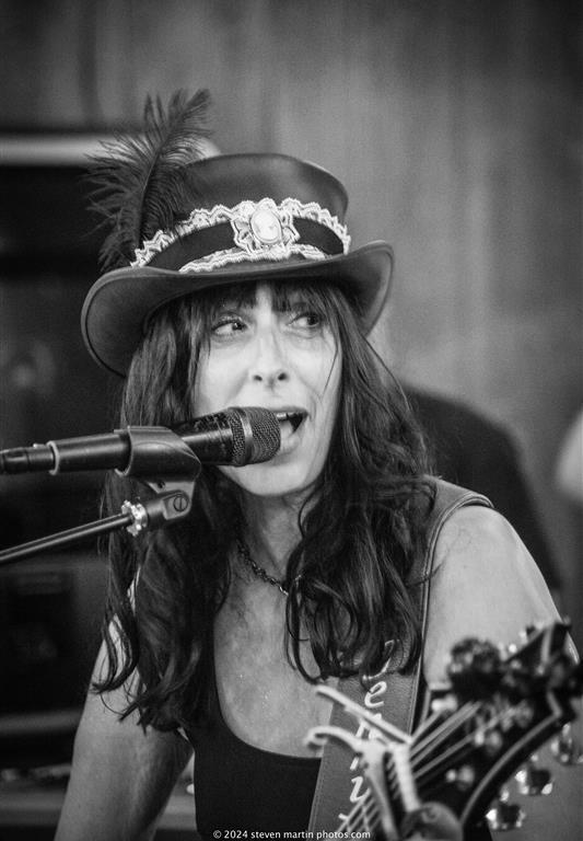 Heather Lynne sings into a microphone while playing guitar, wearing a feathered top hat and dark tank top during a live performance.