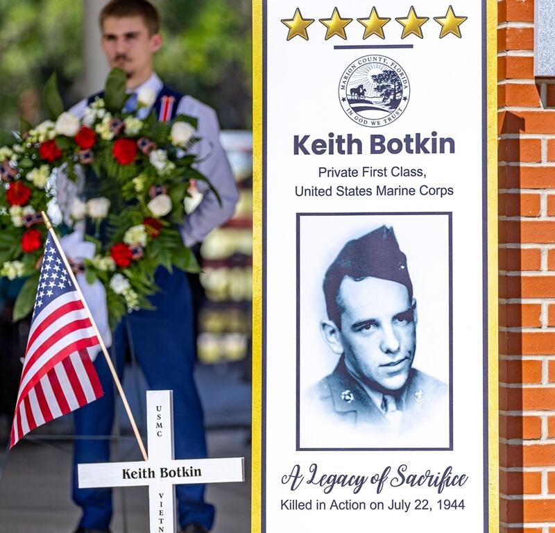 A display honoring USMC Pvt. First Class Keith Botkin features a portrait, a memorial cross, an American flag, and the words “A Legacy of Sacrifice” during a Memorial Day ceremony.