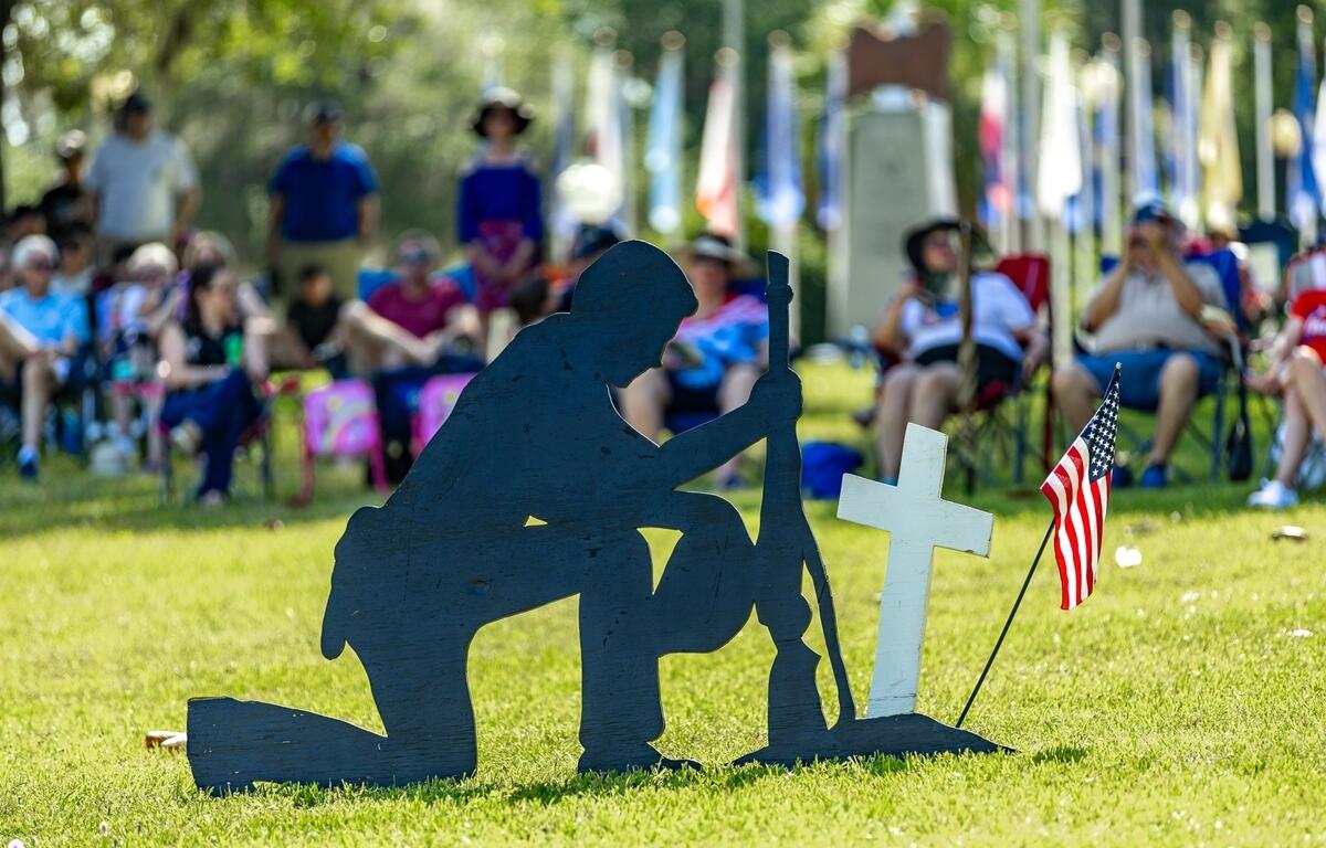 Silhouette of a kneeling soldier with a rifle is placed next to a white cross and American flag in front of a seated crowd at a Memorial Day ceremony.