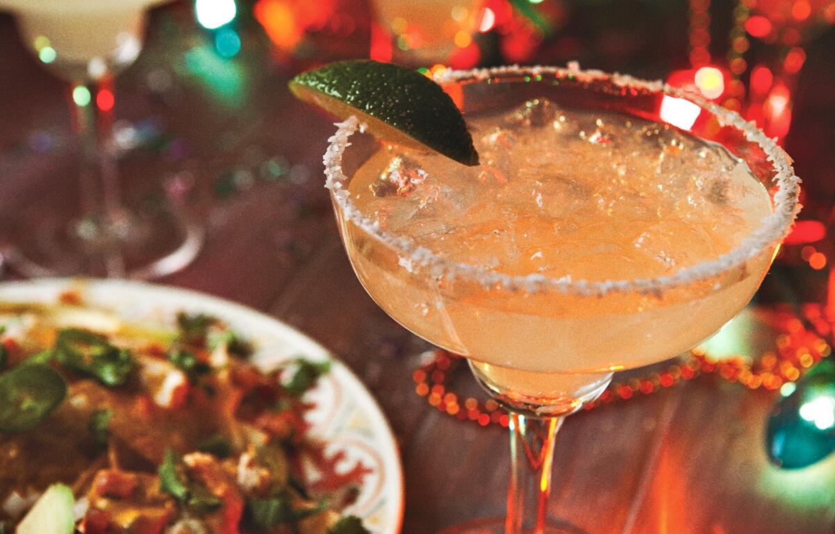 A close-up of a margarita in a glass with a salted rim and lime wedge, next to a colorful plate of Mexican food, with festive string lights in the background.