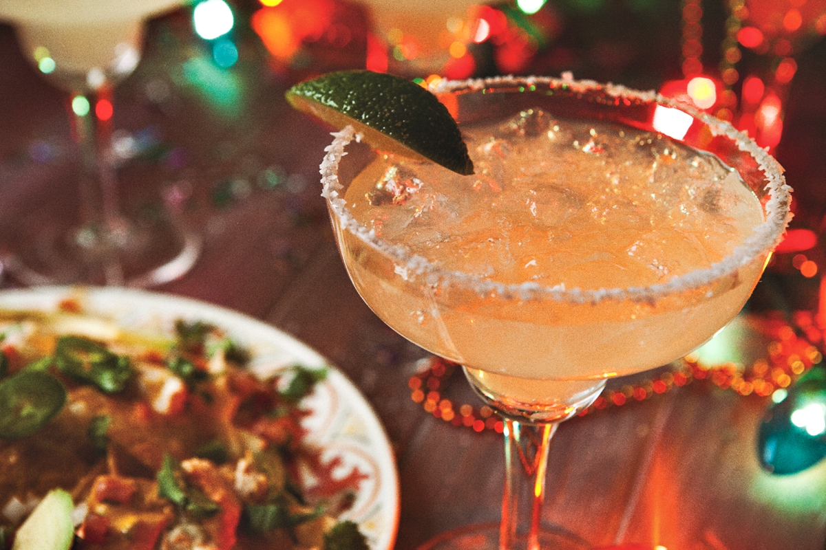 A close-up of a margarita in a glass with a salted rim and lime wedge, next to a colorful plate of Mexican food, with festive string lights in the background.