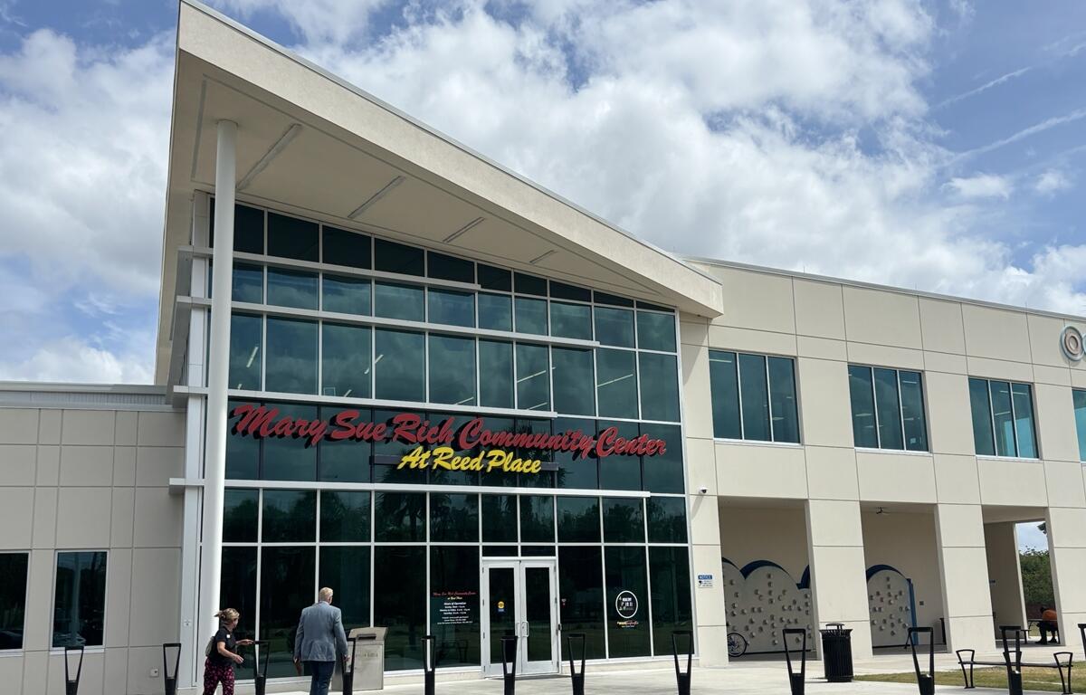Exterior view of the Mary Sue Rich Community Center at Reed Place, a modern two-story building with large glass windows and visitors walking toward the main entrance under a partly cloudy sky.