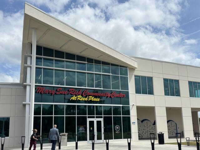 Exterior view of the Mary Sue Rich Community Center at Reed Place, a modern two-story building with large glass windows and visitors walking toward the main entrance under a partly cloudy sky.