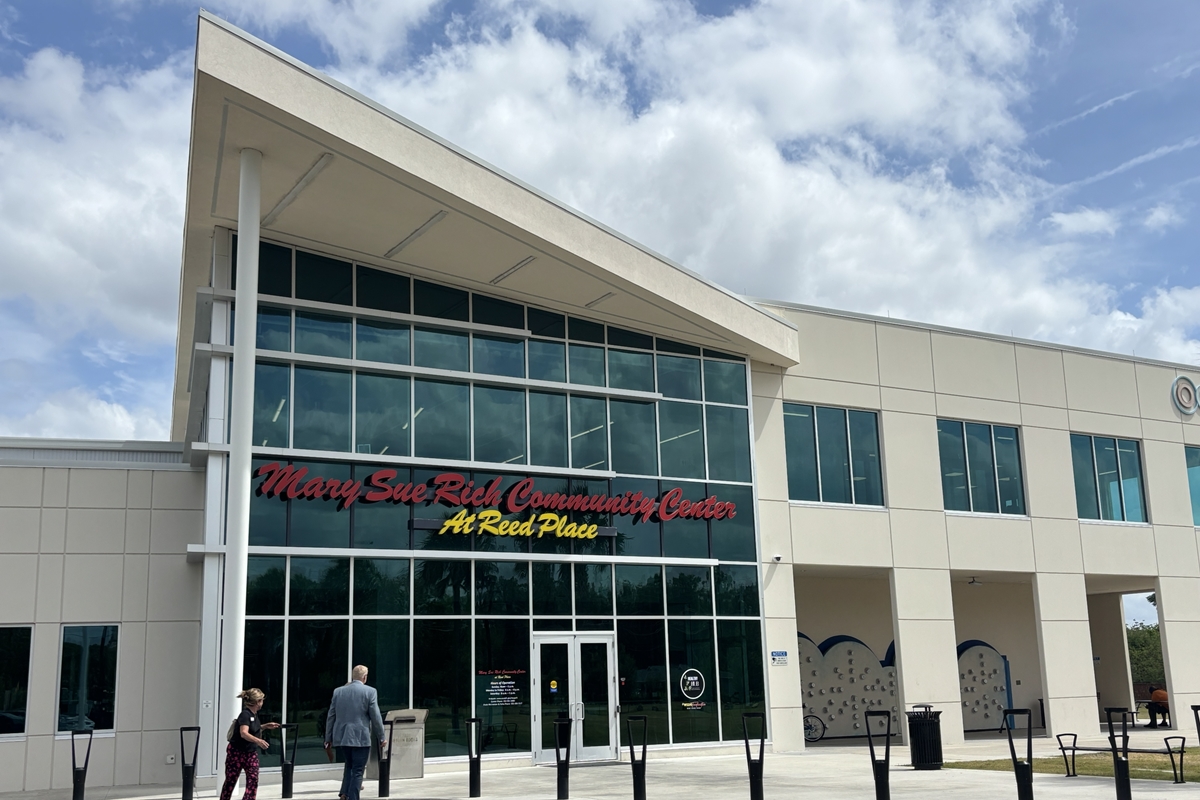 Exterior view of the Mary Sue Rich Community Center at Reed Place, a modern two-story building with large glass windows and visitors walking toward the main entrance under a partly cloudy sky.