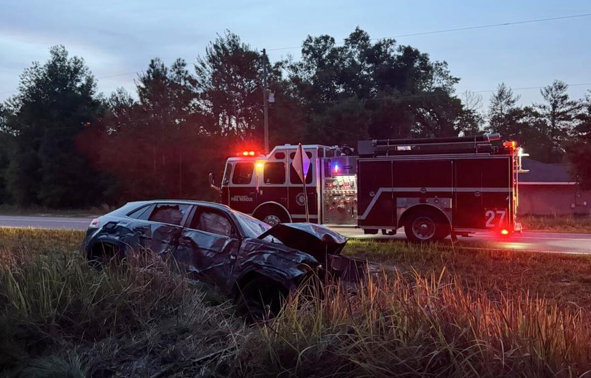 Crashed SUV with heavy front and side damage in a grassy ditch, with Marion County Fire Rescue fire engine parked nearby with emergency lights on.