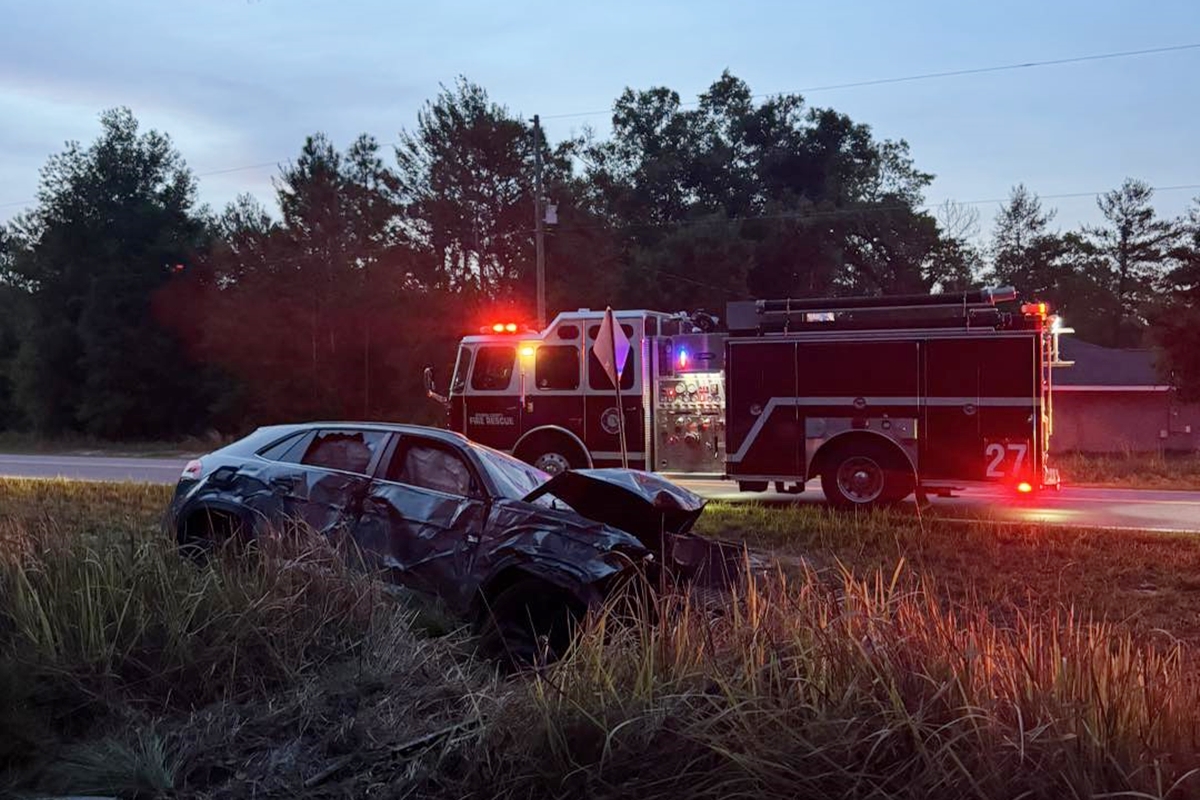 Crashed SUV with heavy front and side damage in a grassy ditch, with Marion County Fire Rescue fire engine parked nearby with emergency lights on.