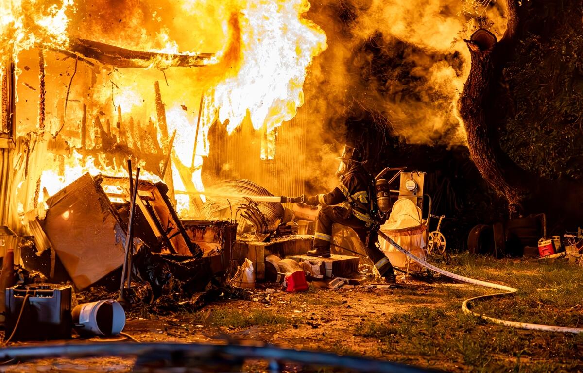 Firefighter sprays water on a fully engulfed mobile home at night, surrounded by flames, smoke, and scattered debris.
