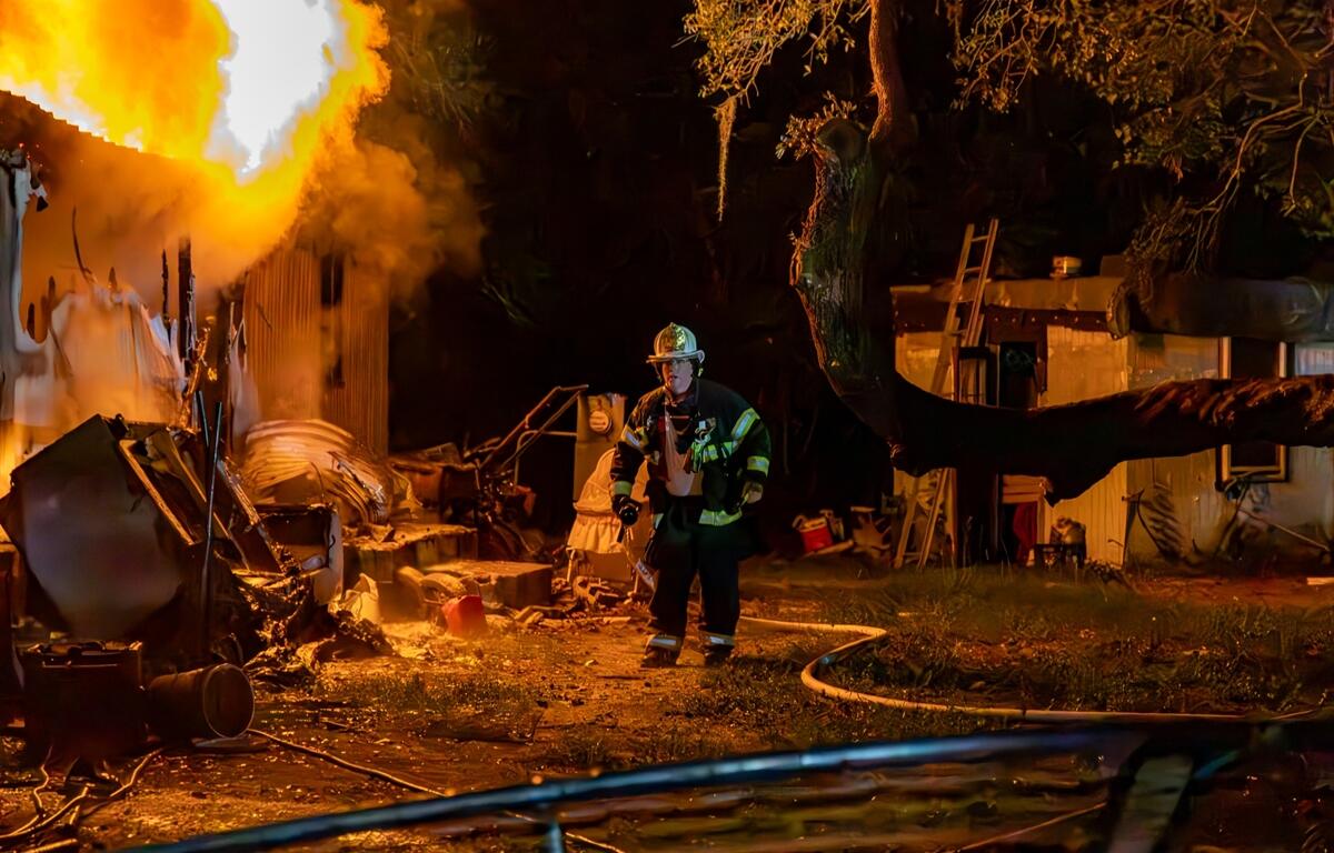Firefighter stands near a burning mobile home at night, surrounded by debris and thick smoke as flames shoot from the structure.